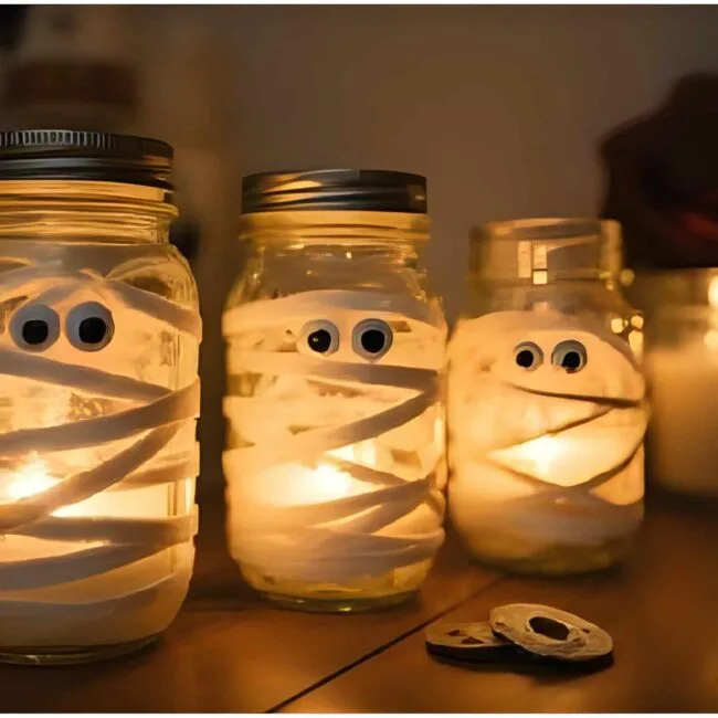 Mason jars wrapped in white gauze with googly eyes peeking out, glowing from tea lights inside. These are displayed on a table with other candlelight and spooky decor - creating a cozy yet eerie vibe.
